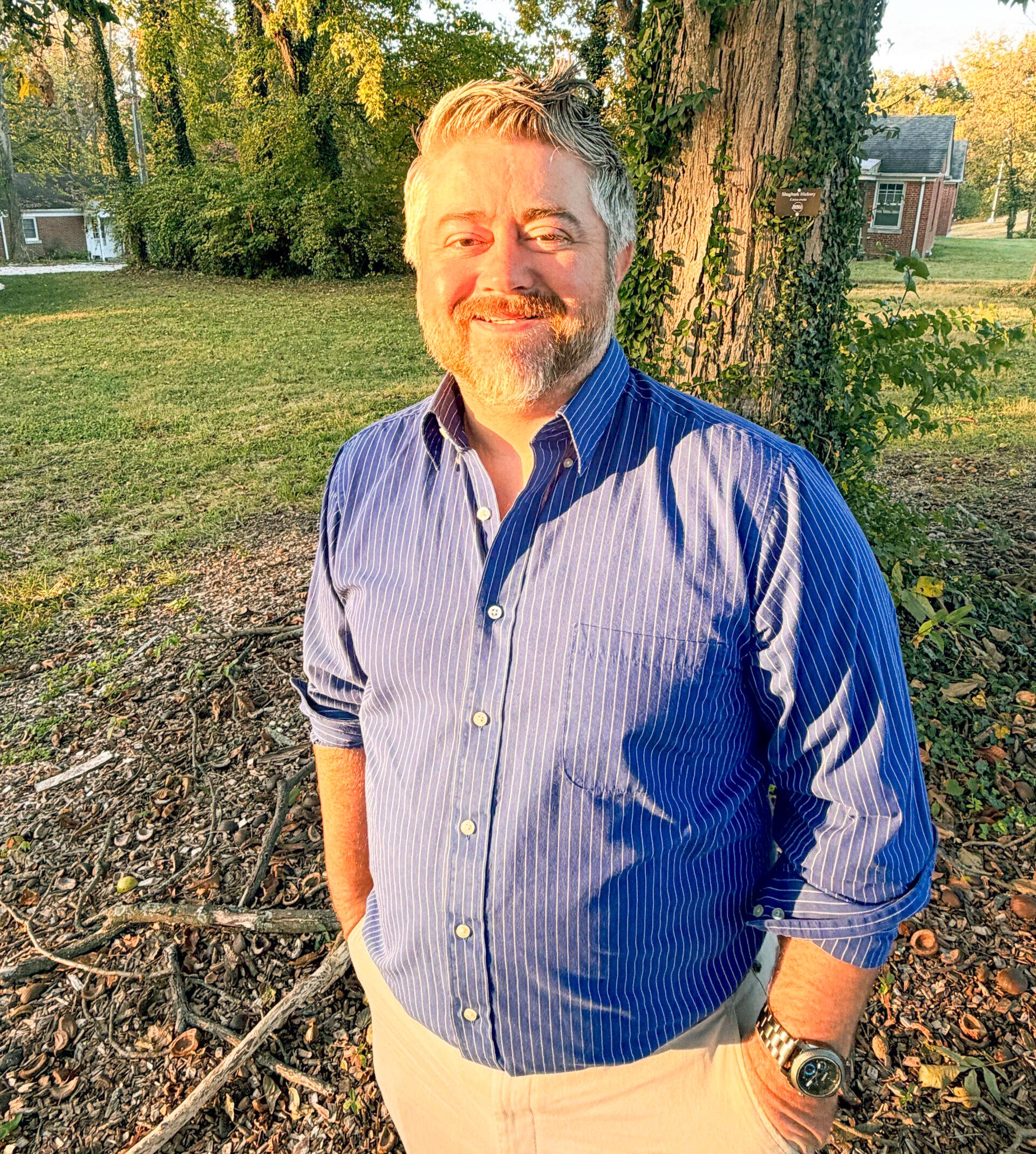 Brandon standing outdoors in front of a tree covered with ivy, wearing a blue button-down shirt.