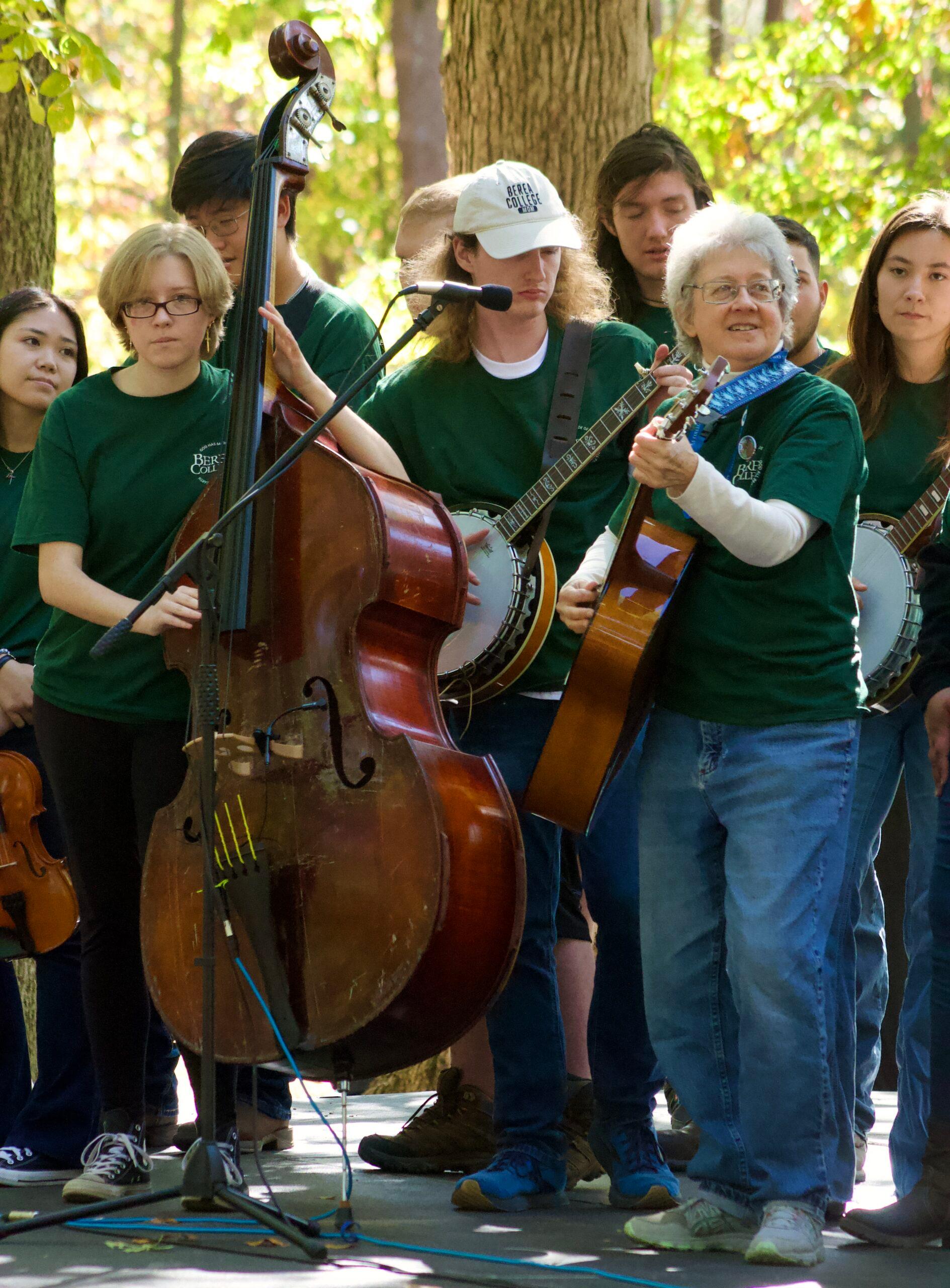 A student ensemble and their director performing outside with guitars, banjos, and upright bass.