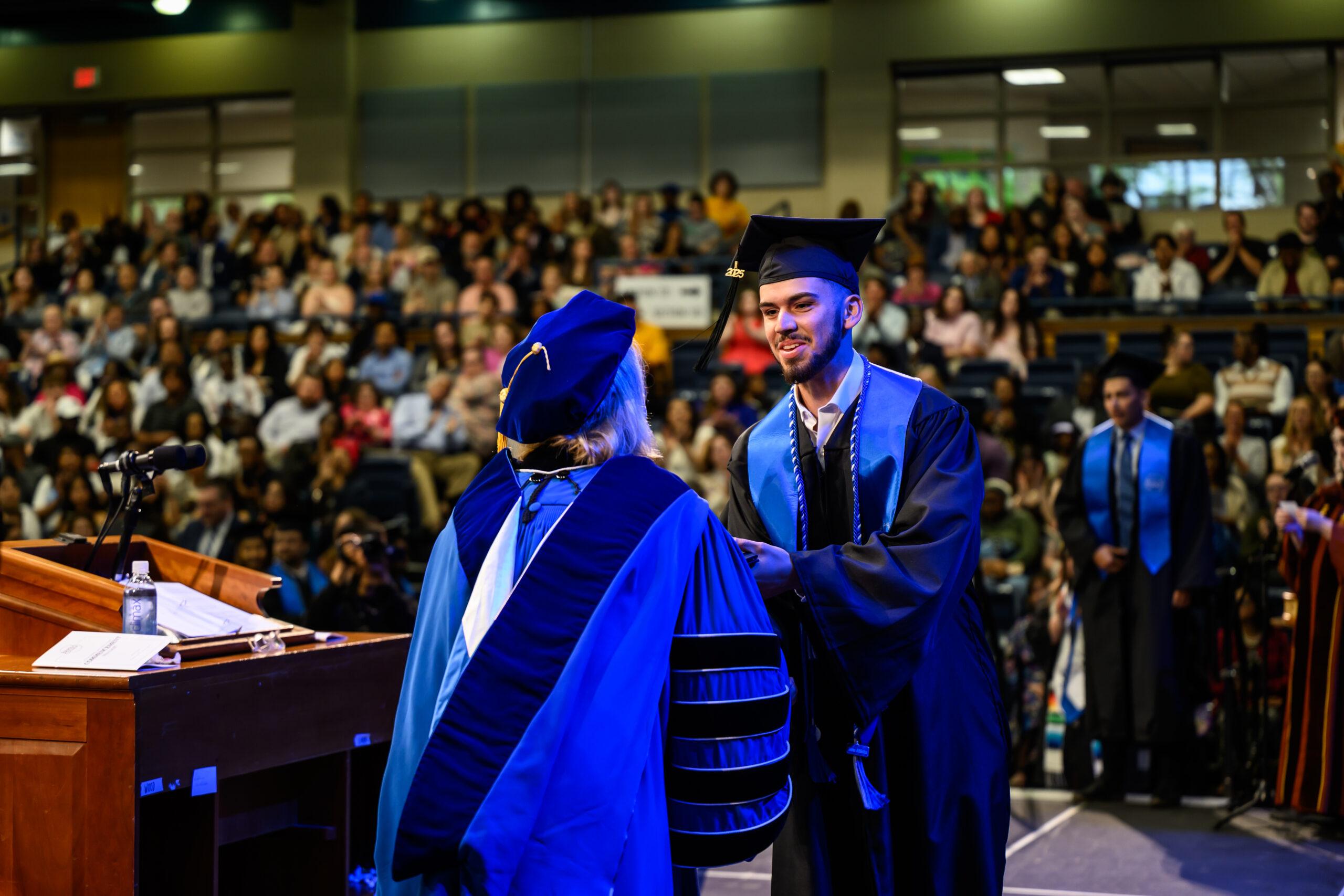 Graduate wearing a cap and gown shakes hands while receiving a diploma on stage at a commencement ceremony, with an audience seated in the auditorium.