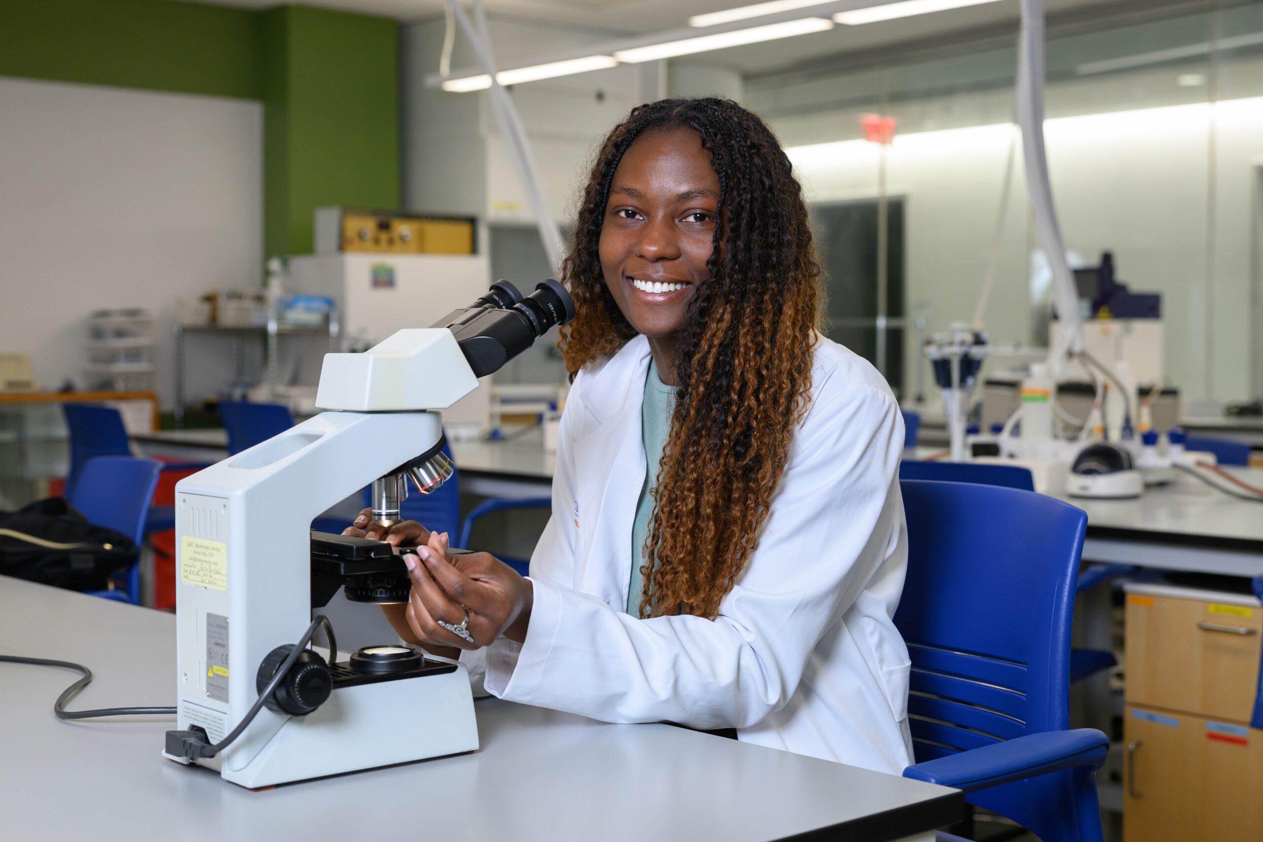 Smiling girl sitting behind a microscope in the MAC Biology lab.
